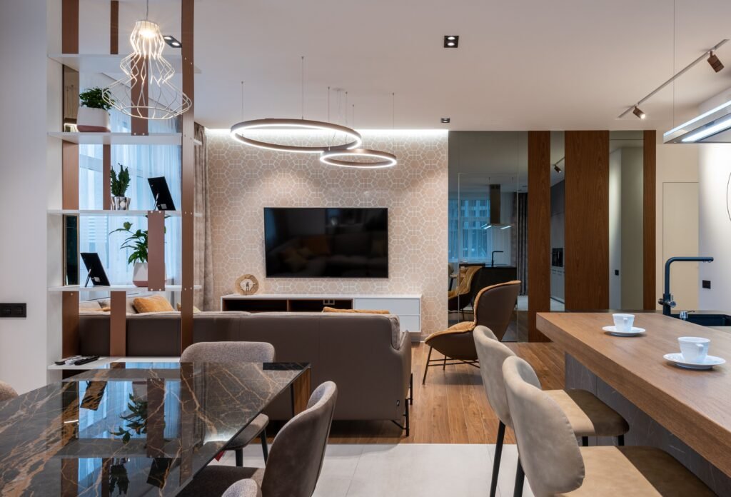 Room with couch and pillows and TV on wall under cabinet near armchair and mirror near window with curtains and shelves near counter with coffee cups and chairs near table